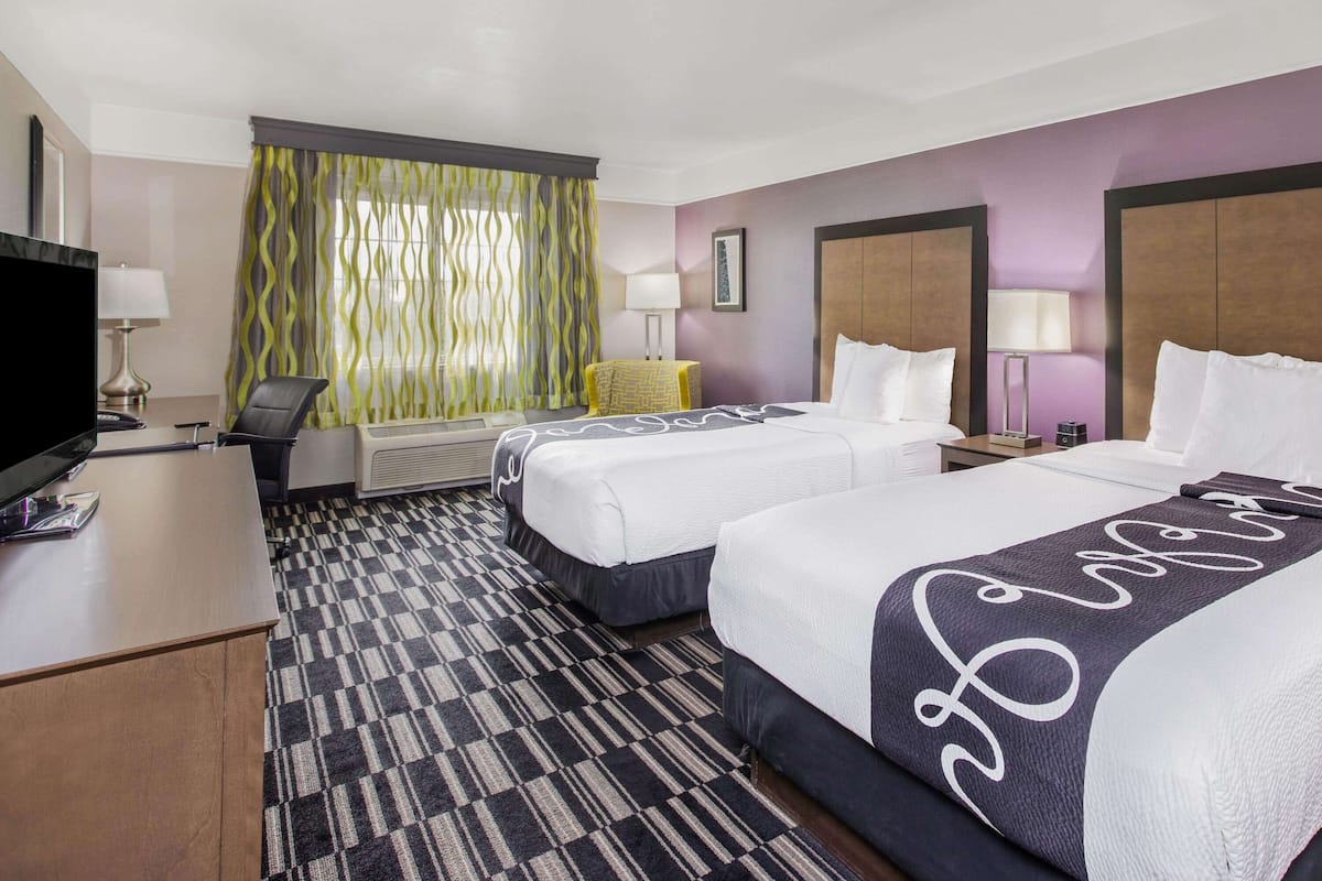 A hotel room with two beds, a desk with a television, a chair, a small table, and a window with curtains.