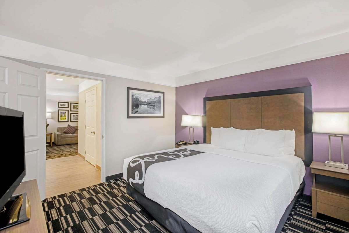 A hotel room with a large bed, a TV, a desk, and a view of a hallway with a sofa and a door.