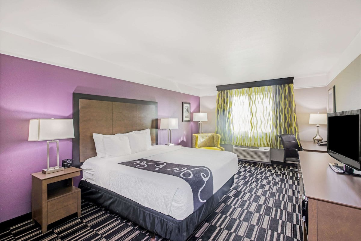 A hotel room with a large bed, a desk with a flat-screen TV, a chair, and a desk lamp.