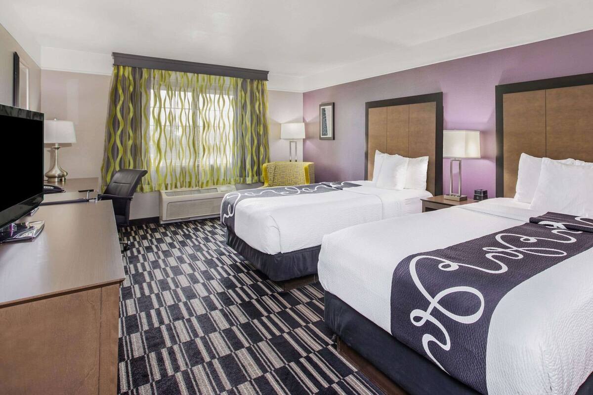 A hotel room with two beds, a desk, a television, and a large window with curtains.