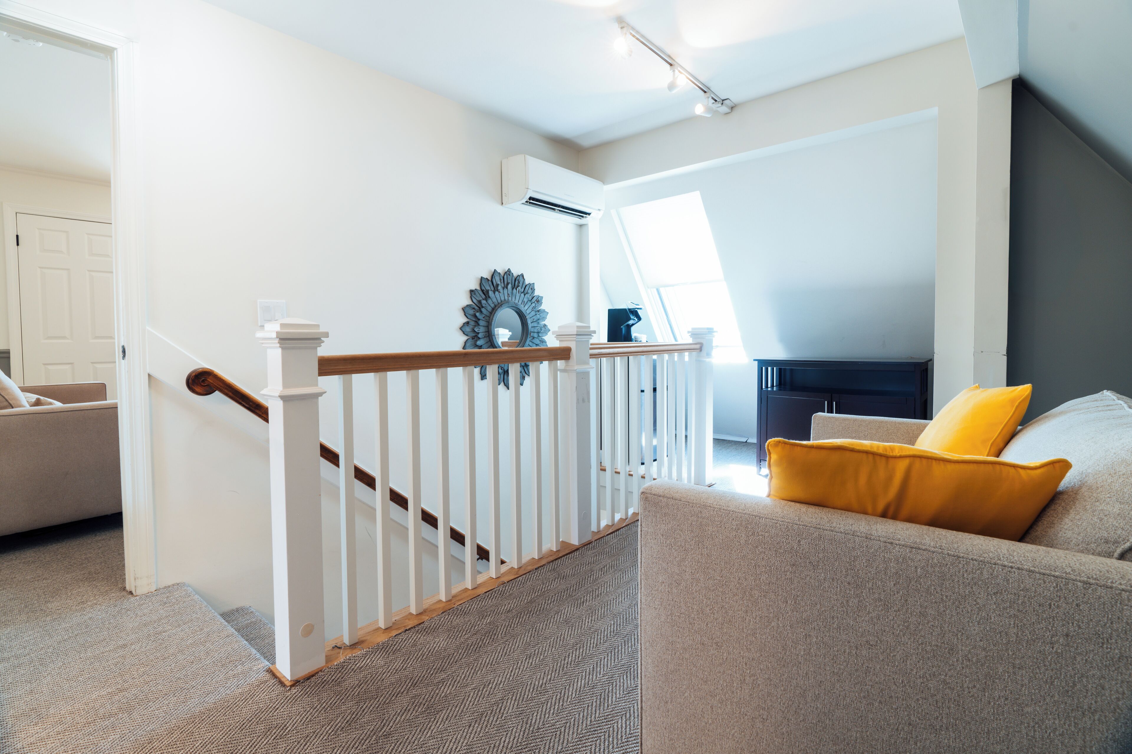 A modern living room with a staircase, a sofa with a yellow pillow, and a decorative mirror.