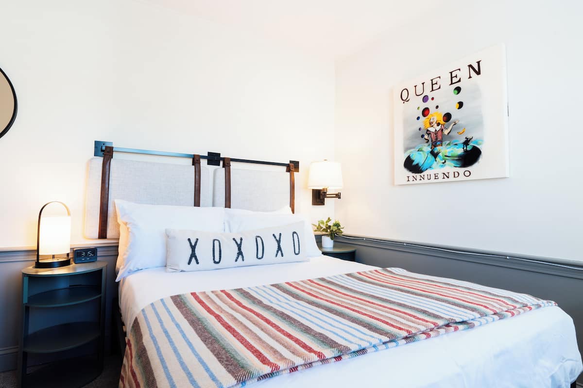 A neatly made bed with a striped blanket, a pillow with "XOXO" text, a wall clock, a bedside lamp, and a poster of a cartoon character.