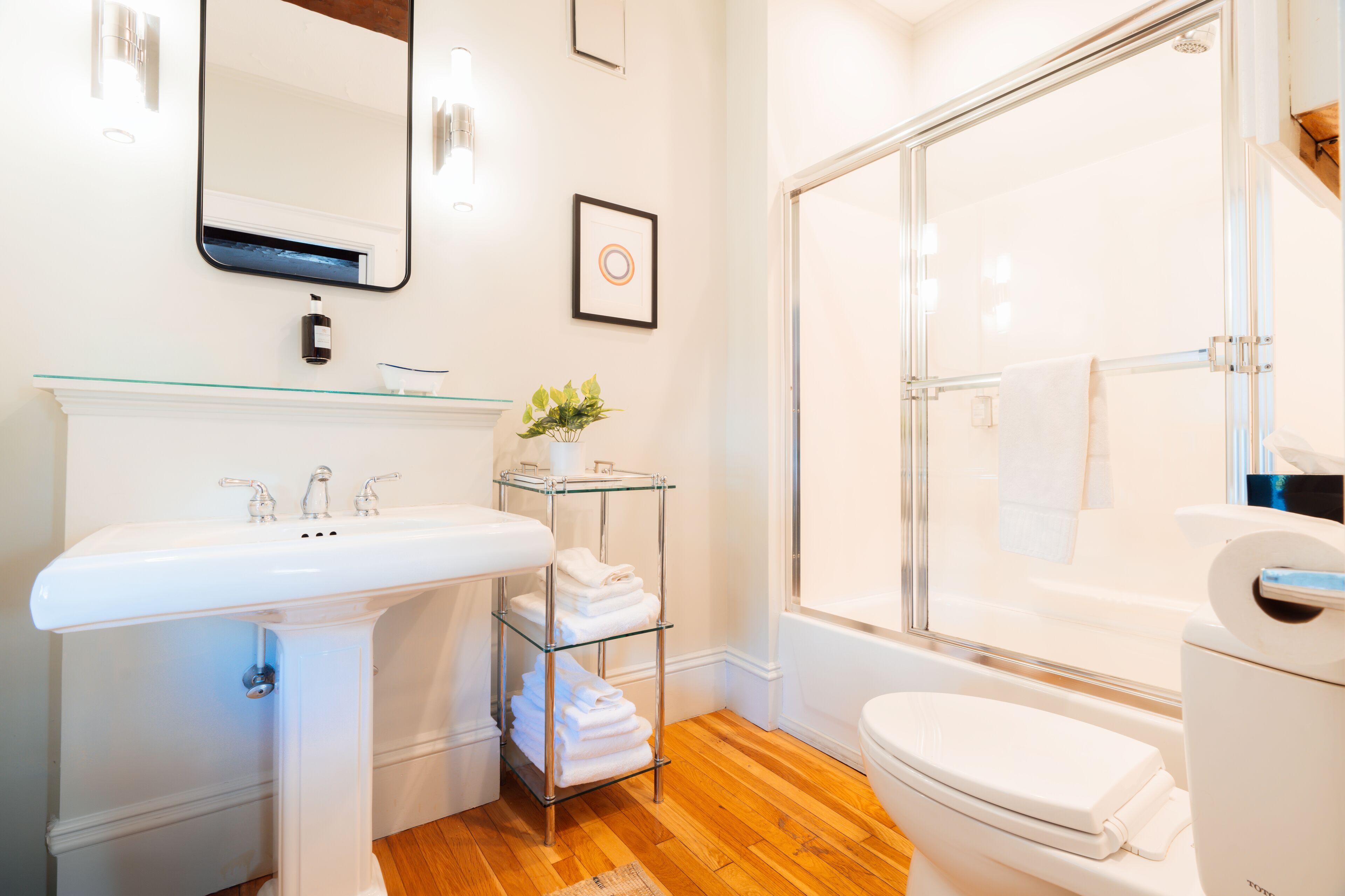 A modern bathroom with a glass shelf holding towels, a white sink, a toilet, and a glass-enclosed shower.