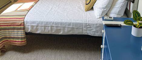A bedroom with a bed, a bedside table with a lamp, a small potted plant, and a window with blinds.