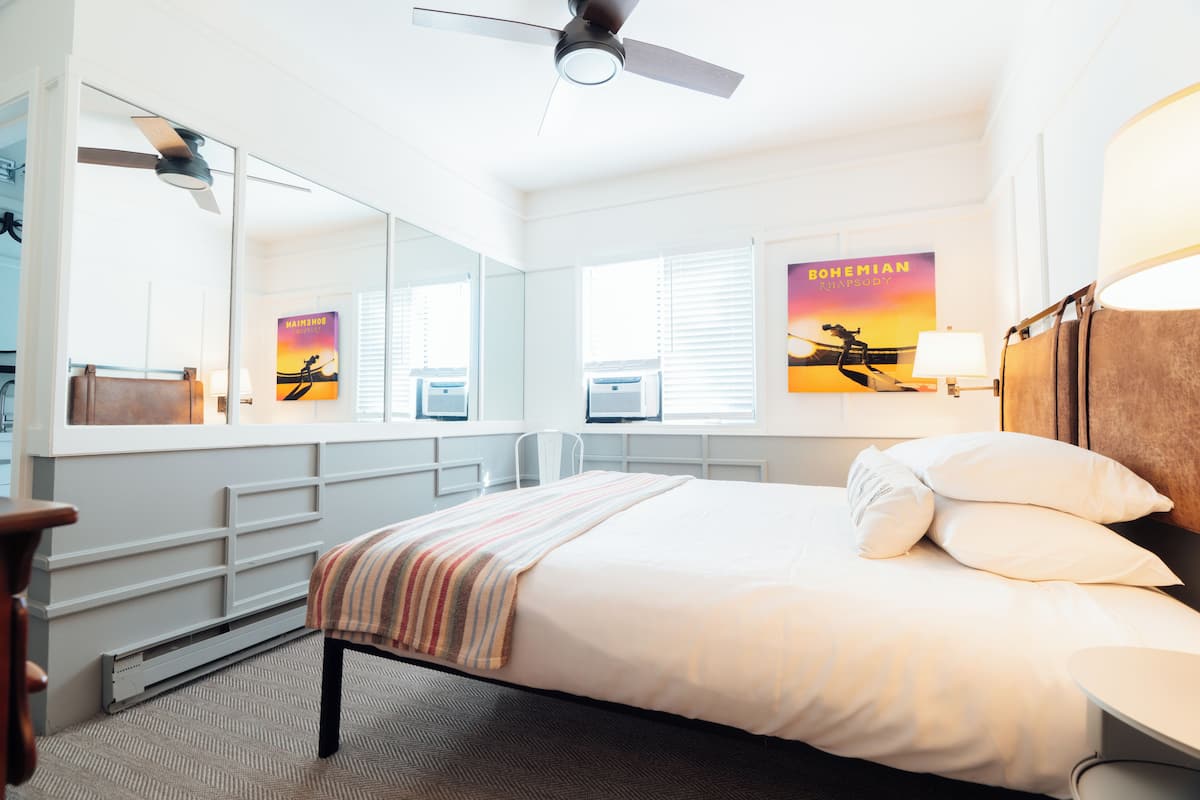 A hotel room with a bed, a desk, a chair, a ceiling fan, a window with blinds, and a poster on the wall.