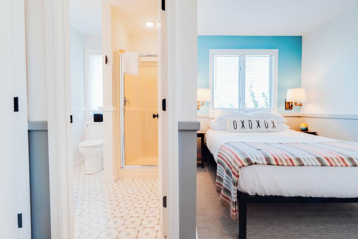 A hotel room with a bed, a window with blinds, a nightstand with a lamp, and a bathroom with a toilet and shower.
