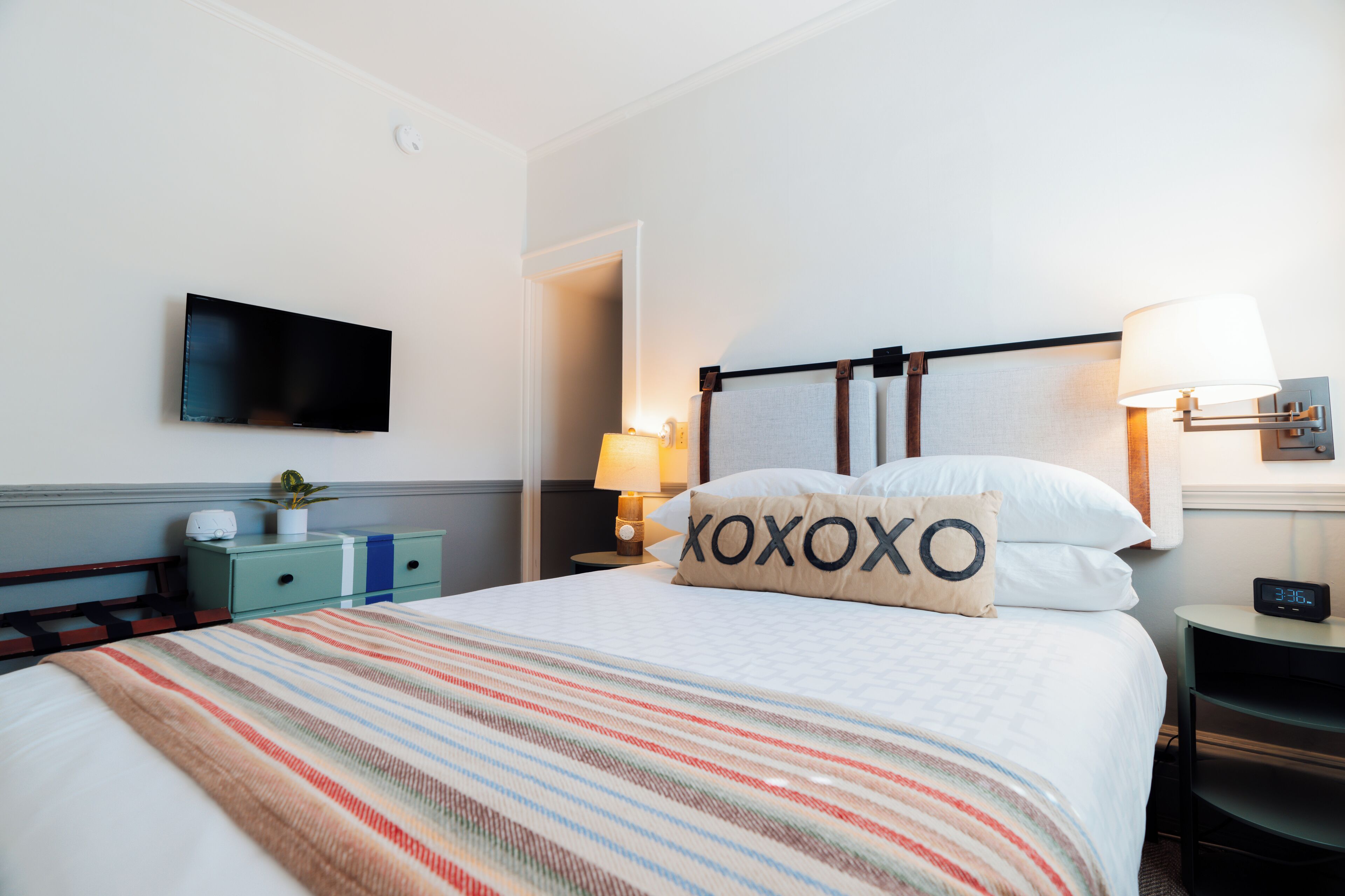 A hotel room with a bed, a TV mounted on the wall, a bedside table with a lamp, and a nightstand with a clock.
