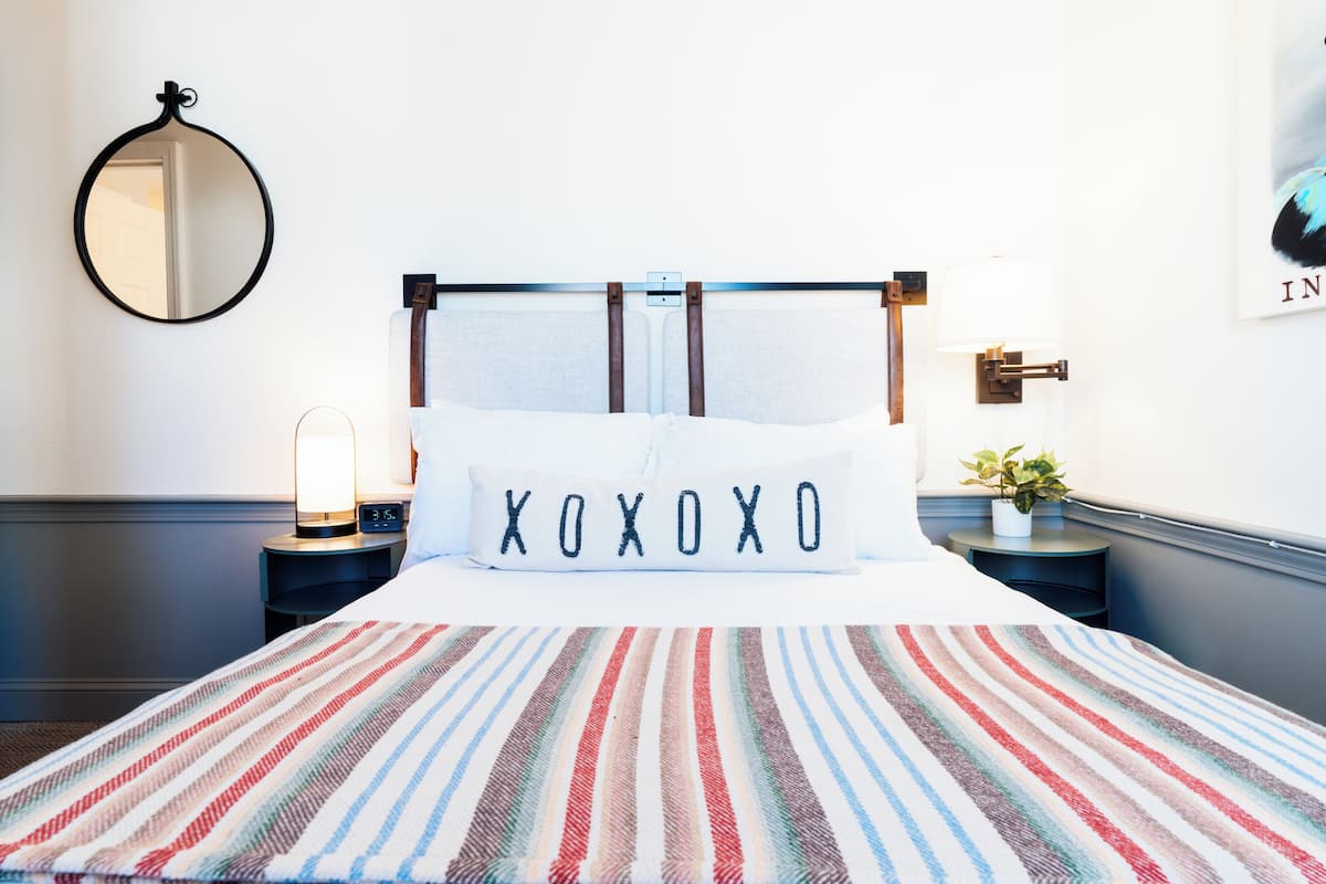 A neatly made bed with a striped blanket, a bedside lamp, a wall-mounted mirror, and a framed picture on the wall.