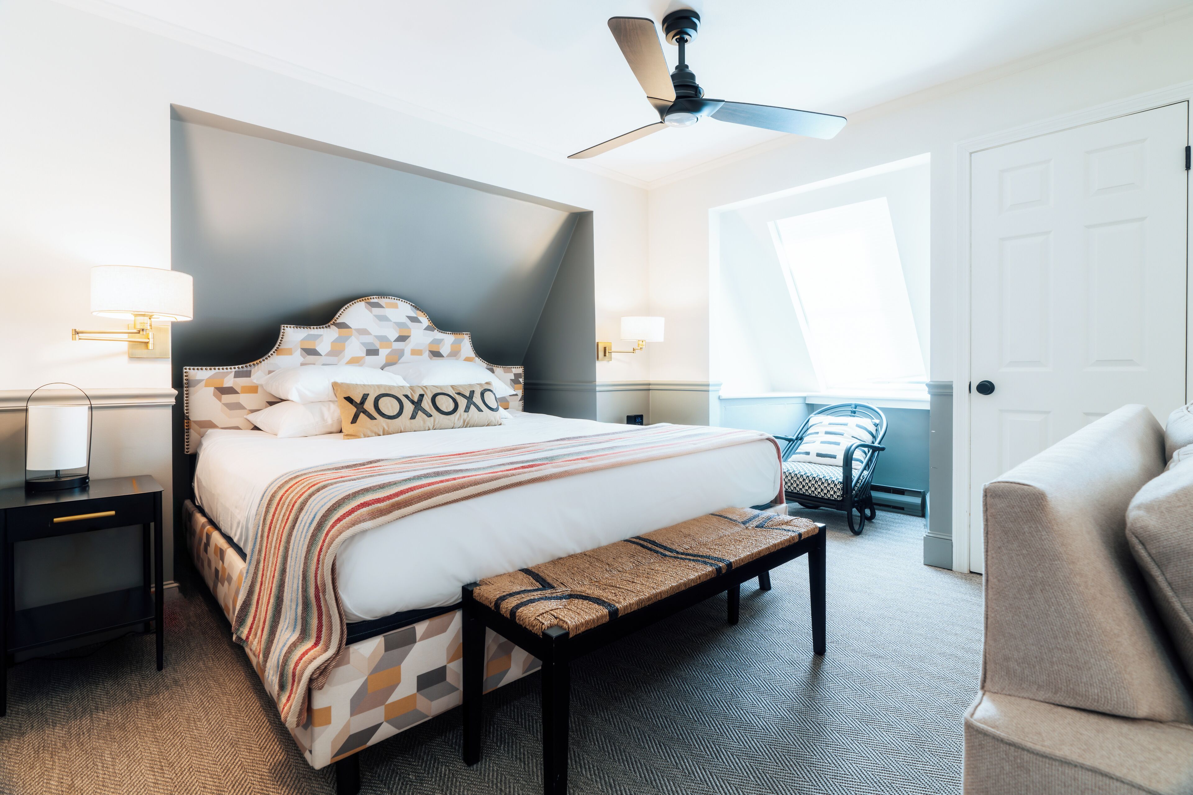 Siististi järjestetty hotellihuone, jossa on suuri sänky, yöpöydällä olevat lamput, penkki, kattotuuletin ja näkymä ulos.