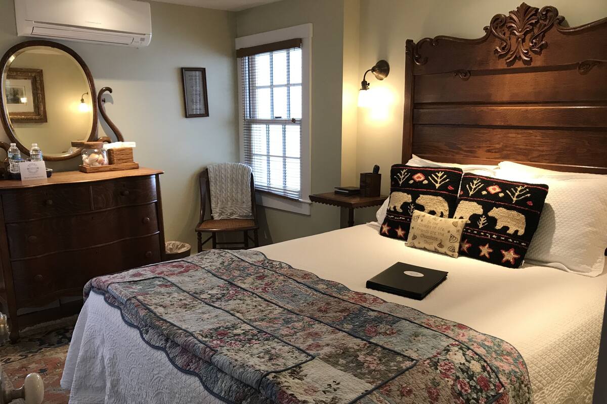 Ein ordentlich gemachtes Bett mit floraler Steppdecke, ein Laptop auf dem Bett, eine hölzerne Kommode, ein Stuhl, ein Fenster mit Jalousien und eine Wandlampe.