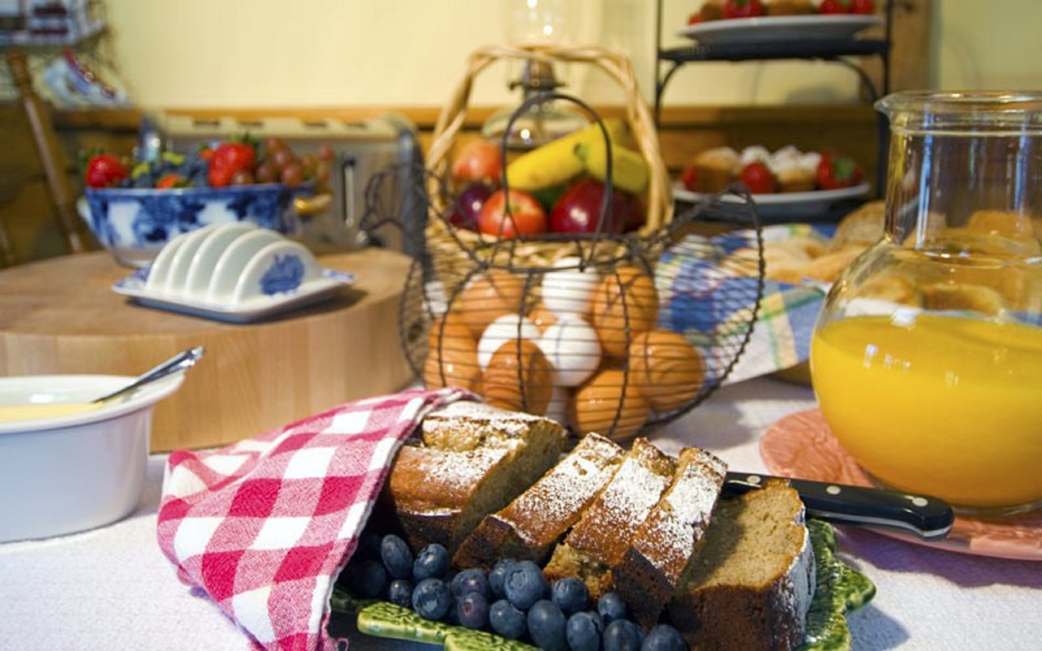 Una mesa de desayuno con rebanadas de pan, arándanos, una cesta de huevos y una jarra de jugo de naranja.