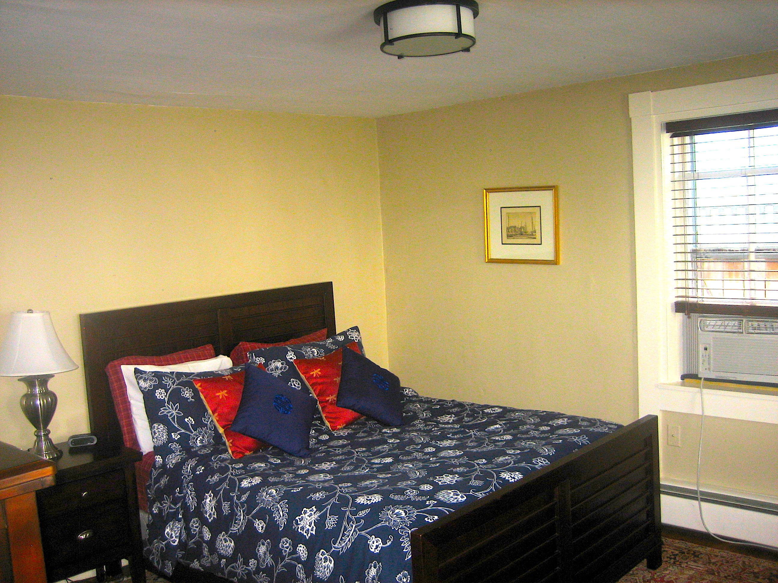 Une chambre à coucher avec un lit, une table de chevet, une lampe, un tableau encadré au mur et une fenêtre avec des stores.