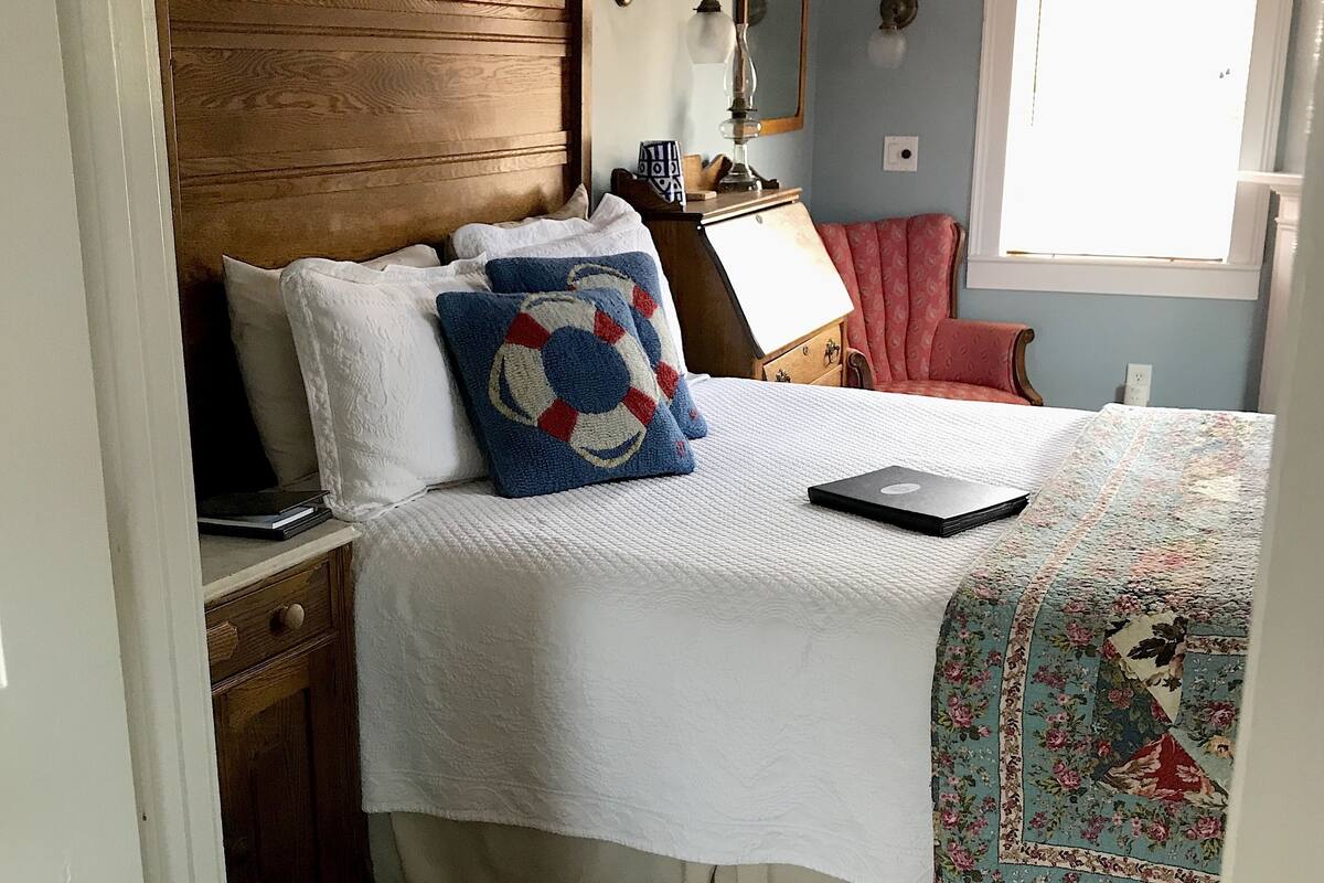 Ein Schlafzimmer mit hölzernem Kopfteil, einem Bett mit weißer Bettwäsche, einem Laptop auf dem Nachttisch, einem roten Sessel und einem Fenster mit Jalousien.