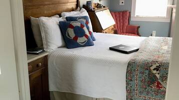 Quarto com cabeceira de madeira, cama com roupa de cama branca, um laptop sobre a mesa de cabeceira, uma poltrona vermelha e uma janela com persianas.