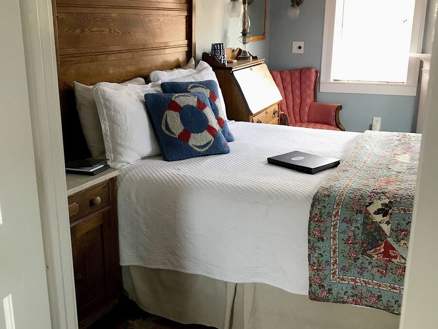 Quarto com cabeceira de madeira, cama com roupa de cama branca, um laptop sobre a mesa de cabeceira, uma poltrona vermelha e uma janela com persianas.