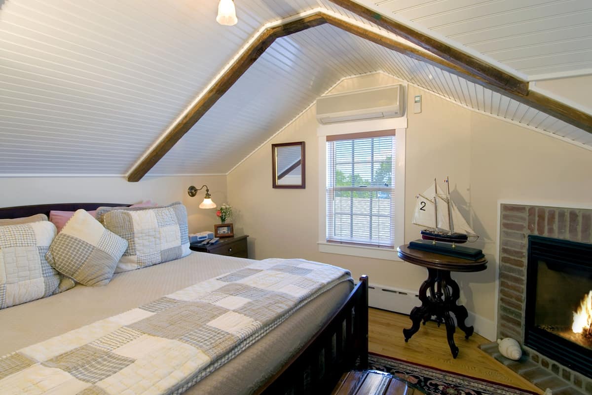 Ein Schlafzimmer mit geneigter Decke, einem Bett mit Bettdecke, einem Nachttisch, einem kleinen Tisch mit Lampe, einem Fenster mit Jalousien und einem Kamin.
