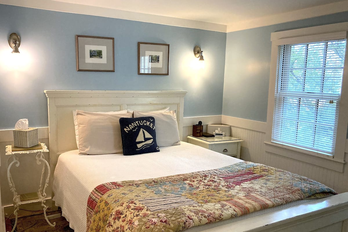 Ein Schlafzimmer mit einem weißen Bett, einer floralen Bettdecke, einem Kissen im Nantucket-Stil, zwei gerahmten Bildern, einem Nachttisch, einer Lampe und einem Fenster mit Jalousien.