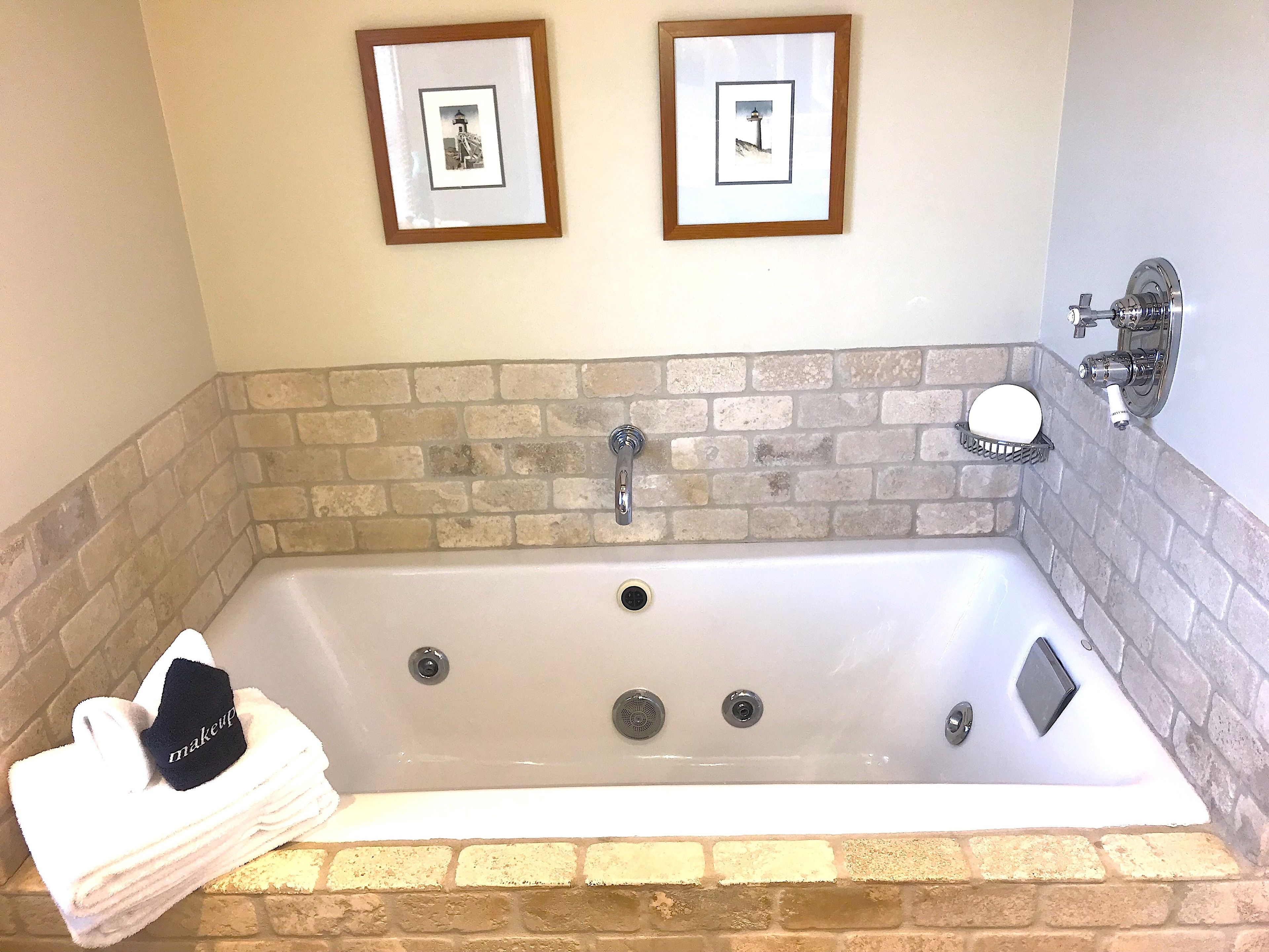 A bathroom with a brick wall, a white bathtub, a towel stack, and two framed pictures on the wall.