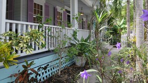 A covered porch with a white railing, a blue bench, and various potted plants and flowers. - The Inn on Fleming (Key West)
