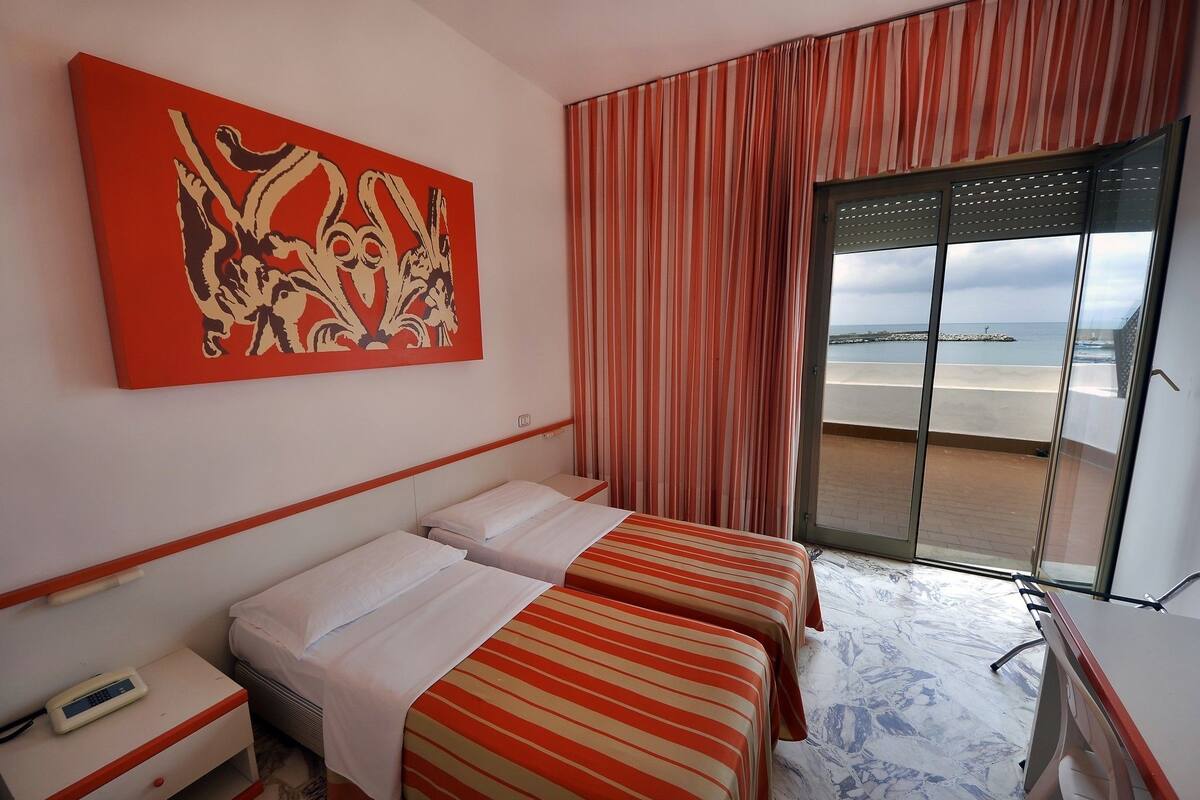 A hotel room with two beds, a painting on the wall, a desk, and a view of the beach through the window.