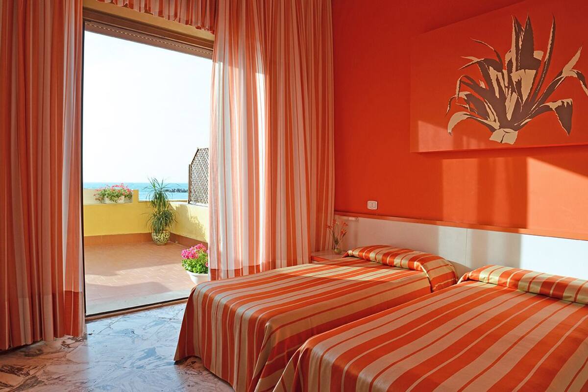 A room with two beds, a balcony with a view, and a large plant on the wall.
