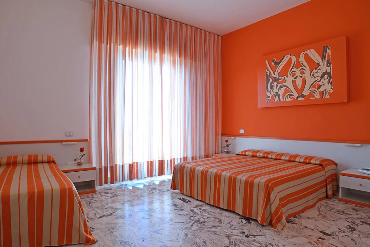 A hotel room with two beds, striped bedding, a marble floor, a large window with curtains, and an orange accent wall with a painting.