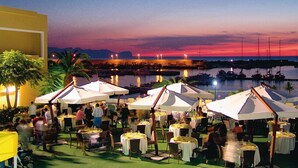 An outdoor dining area with tables and umbrellas, overlooking a marina with boats and a sunset. - Palm Beach Hotel (Cinisi)