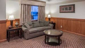 A hotel room with a grey sofa, two side tables with lamps, a round coffee table, a patterned carpet, a window with curtains, and a framed picture on the wall. - Super 8 by Wyndham St Robert Ft Leonard Wood Area (St. Robert)
