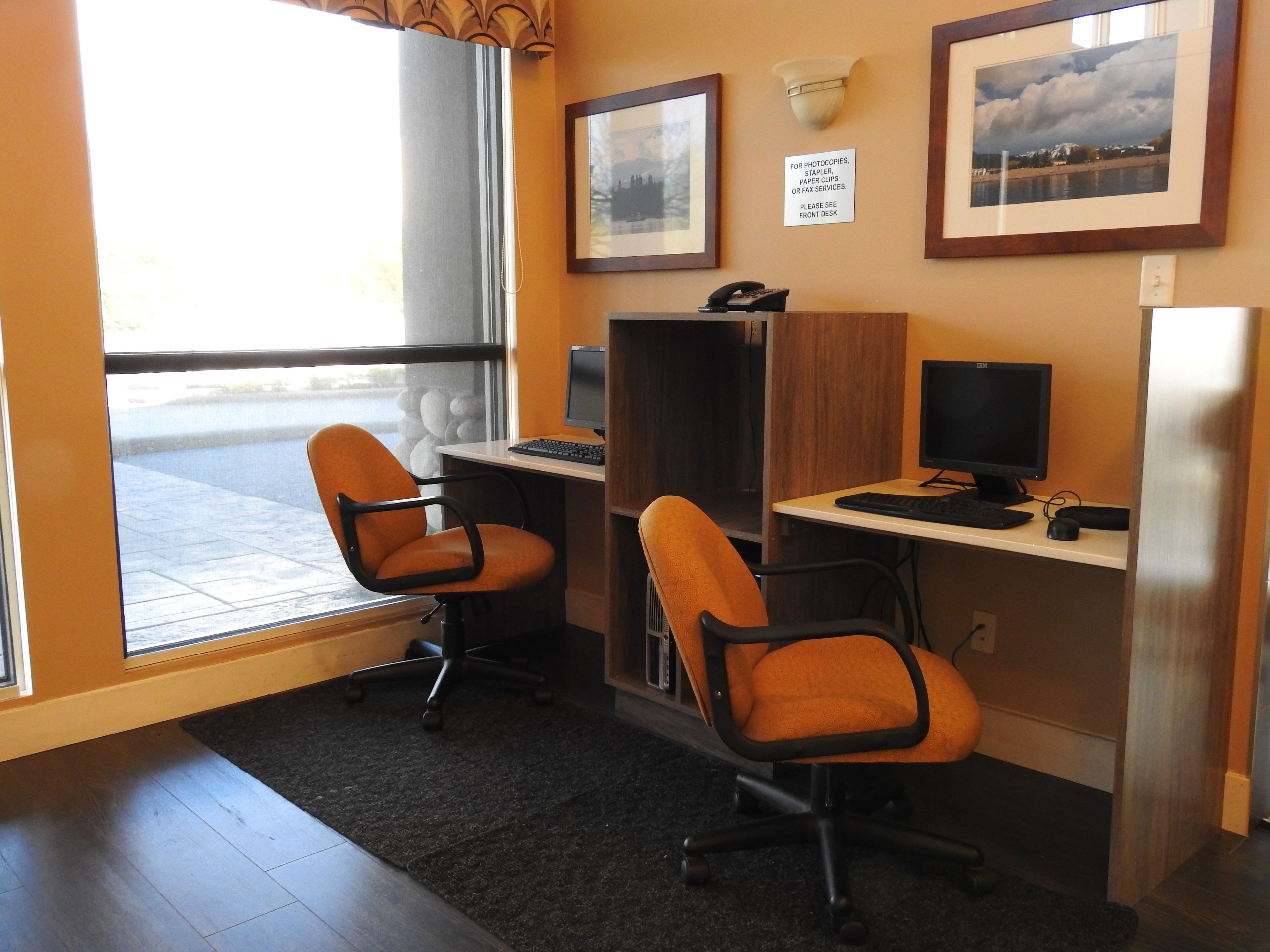 A room with two orange office chairs, two desks with computers, a window with a view of a body of water, and framed pictures on the wall.