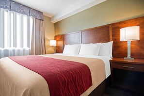 Habitación de hotel con una cama grande, cabecera de madera, mesita de noche con lámpara y ventana con cortinas.