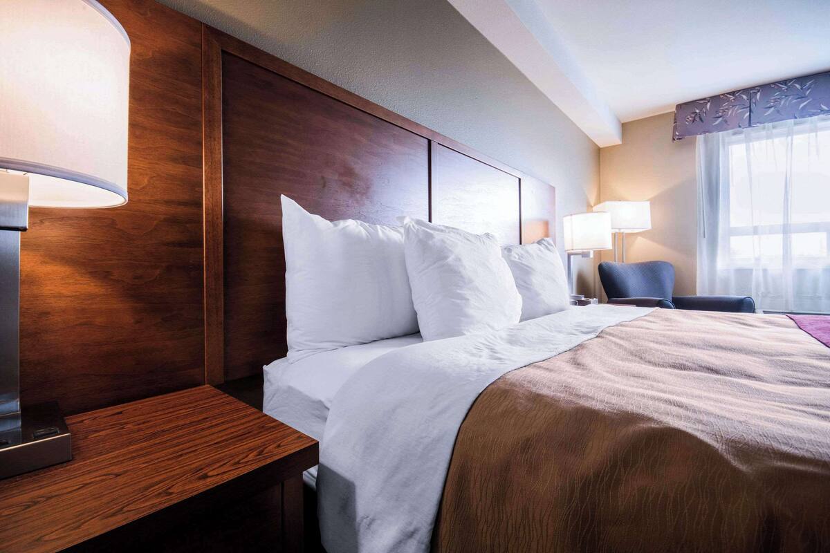 A hotel room with a large bed, wooden headboard, two bedside lamps, a nightstand, and a chair.