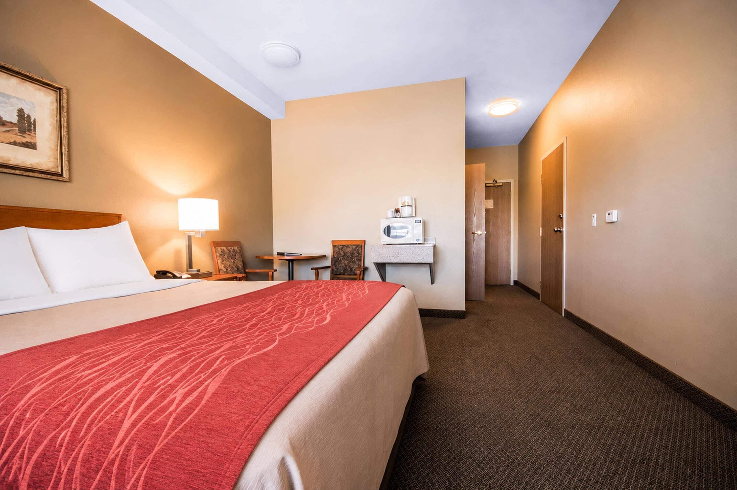 Quarto de hotel com cama, escrivaninha com luminária, cadeira, micro-ondas e porta que dá para outro cômodo.