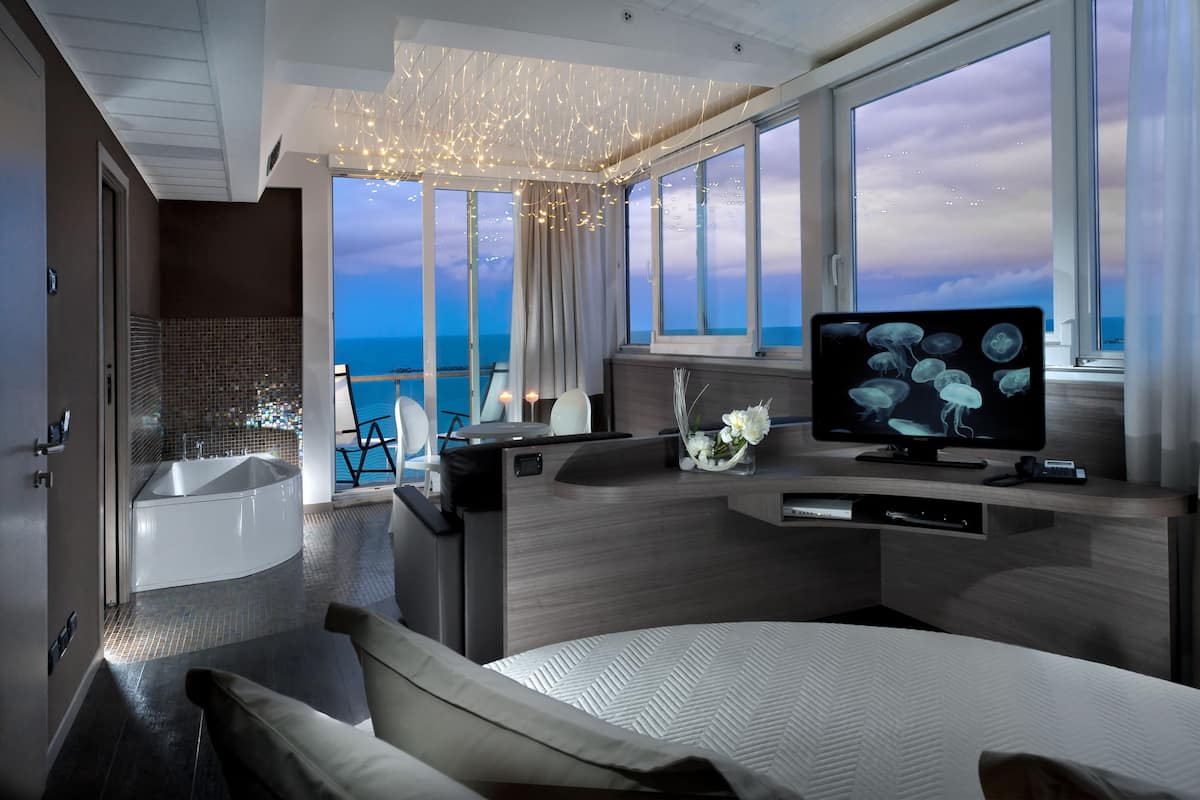 A modern room with a large window, a flat-screen TV, a bathtub, and a seating area with a view of the sea.