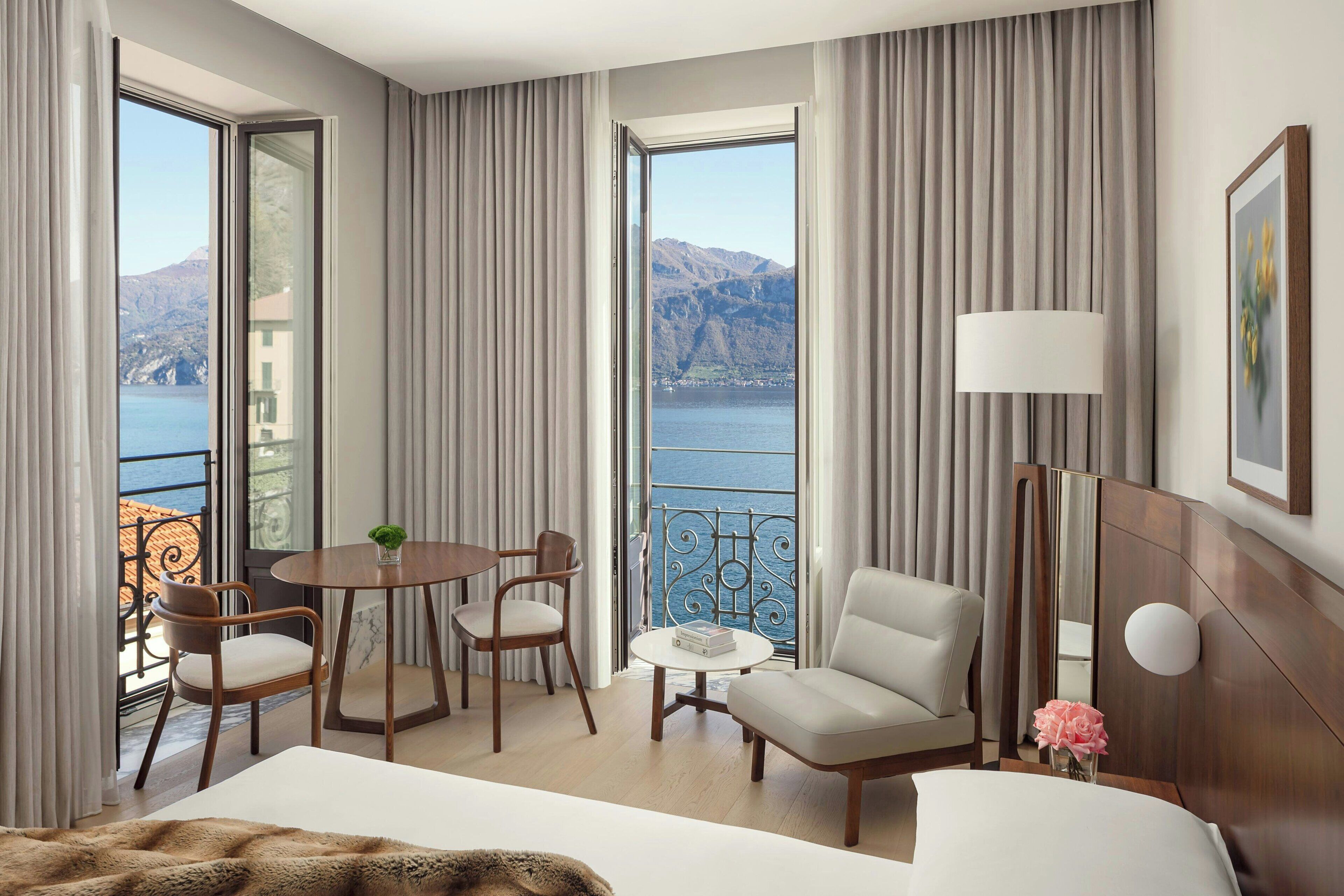 A hotel room with a large bed, a sitting area with a table and chairs, a lamp, and a view of the sea and mountains.