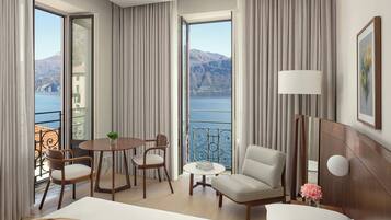 Camera d'albergo con un ampio letto, un'area salotto con tavolo e sedie, una lampada e vista sul mare e sulle montagne.