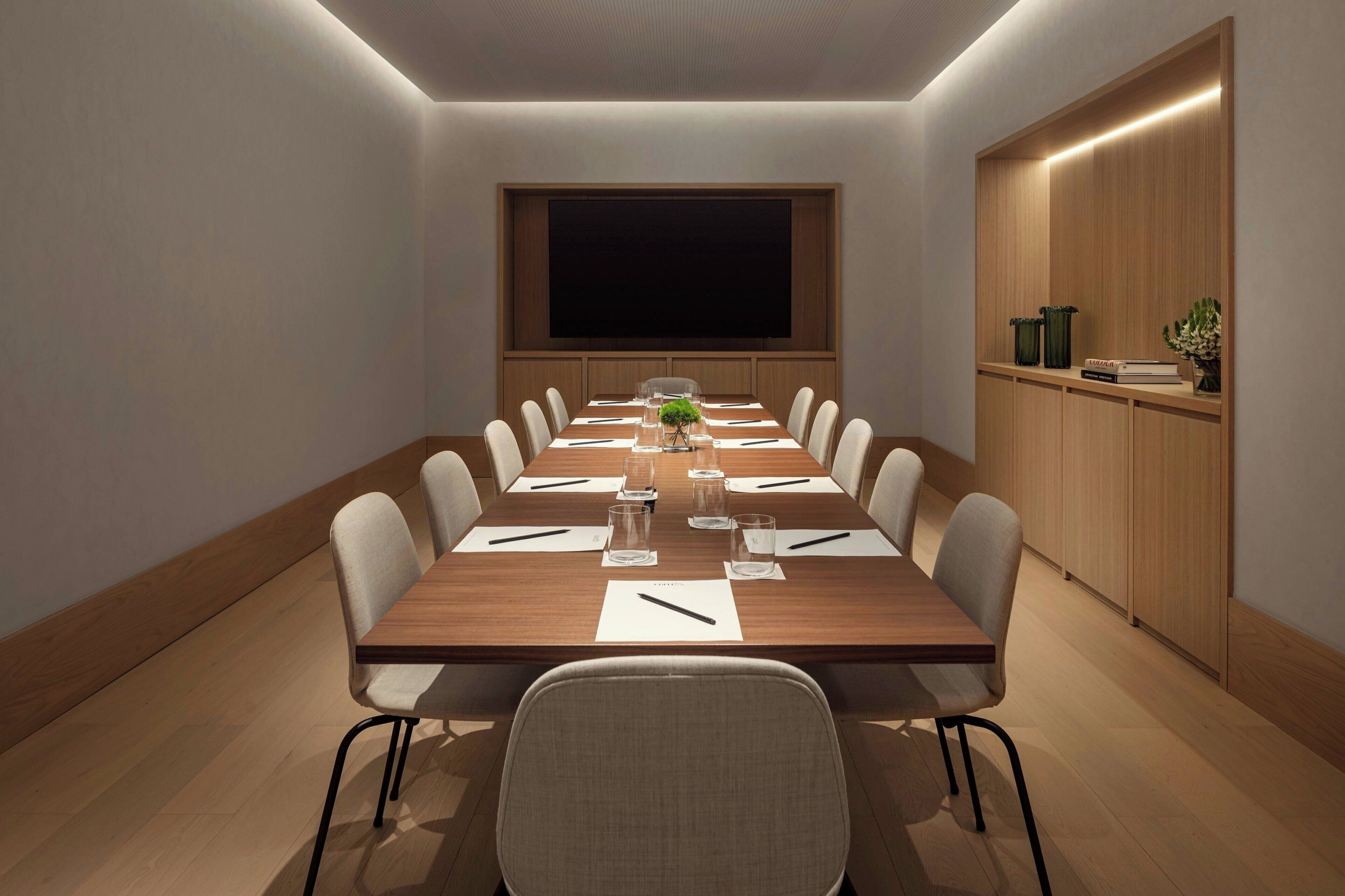 A conference room with a long table, chairs, a projector screen, and a wooden cabinet with books and decorative items.