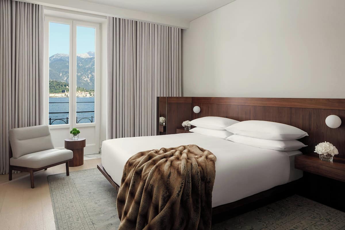 A hotel room with a large bed, a chair, a small table with a vase, and a view of the sea and mountains.