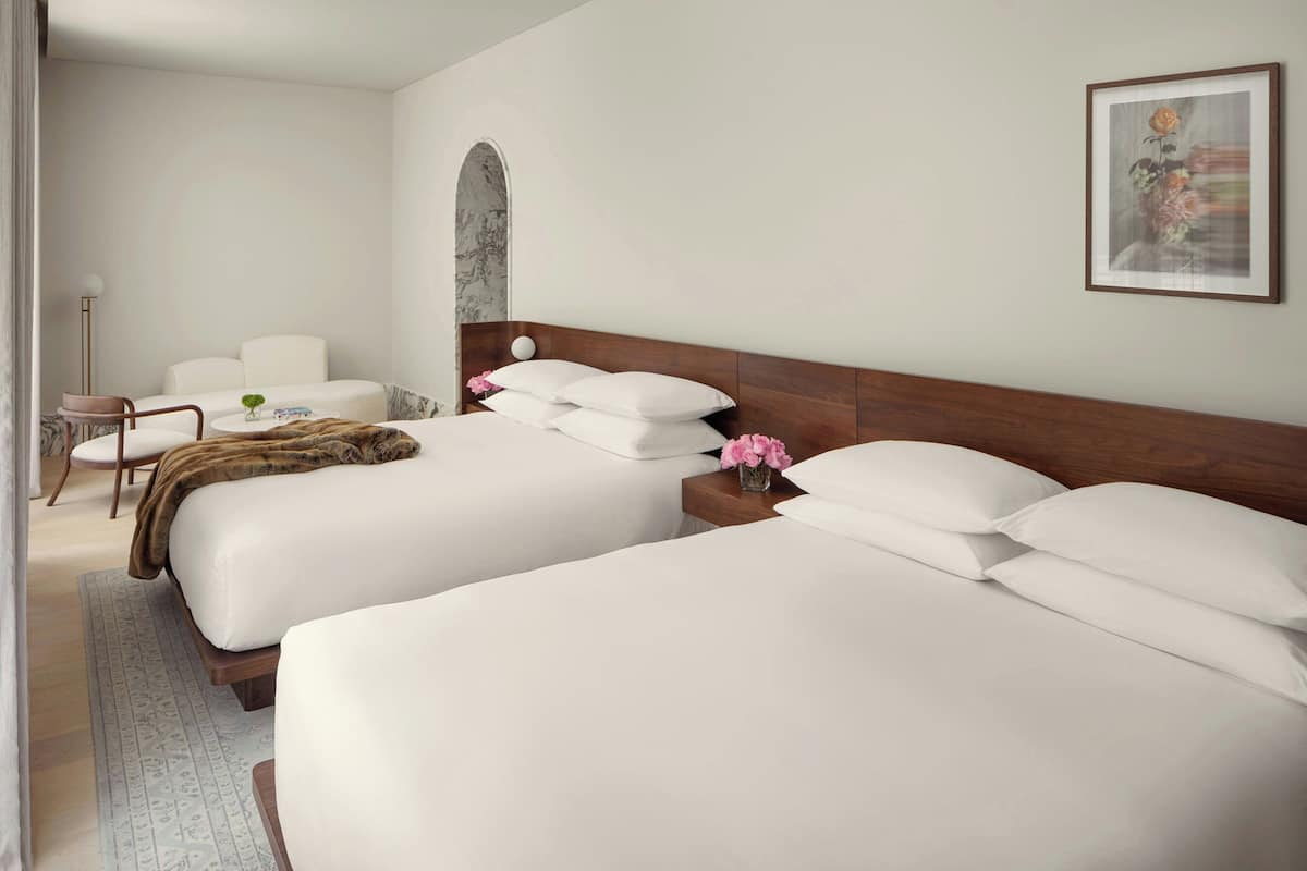 A hotel room with two large beds, a sitting area with a chair and a small table, a framed artwork on the wall, and a decorative arch.