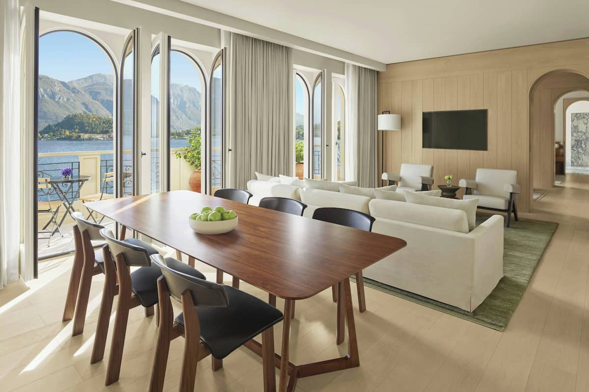 A spacious room with a dining table, chairs, a sofa, and a TV. Large windows offer a view of a lake and mountains.