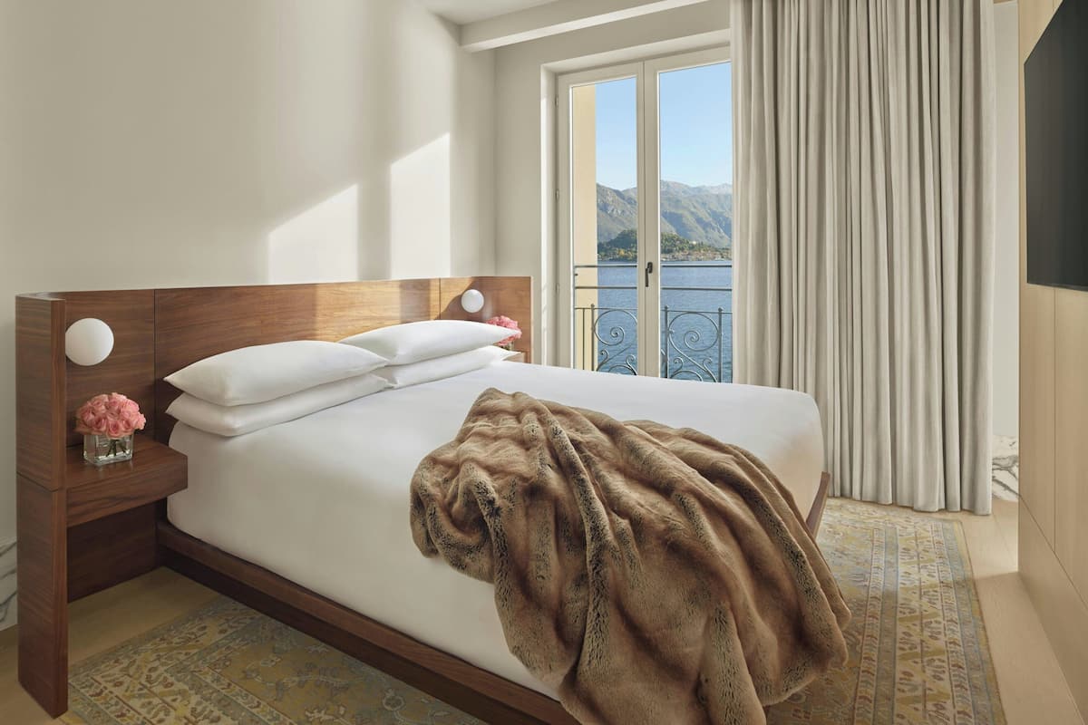 A hotel room with a wooden headboard, a large bed with white linens, a brown throw blanket, a nightstand with a vase of pink flowers, and a view of mountains through the window.