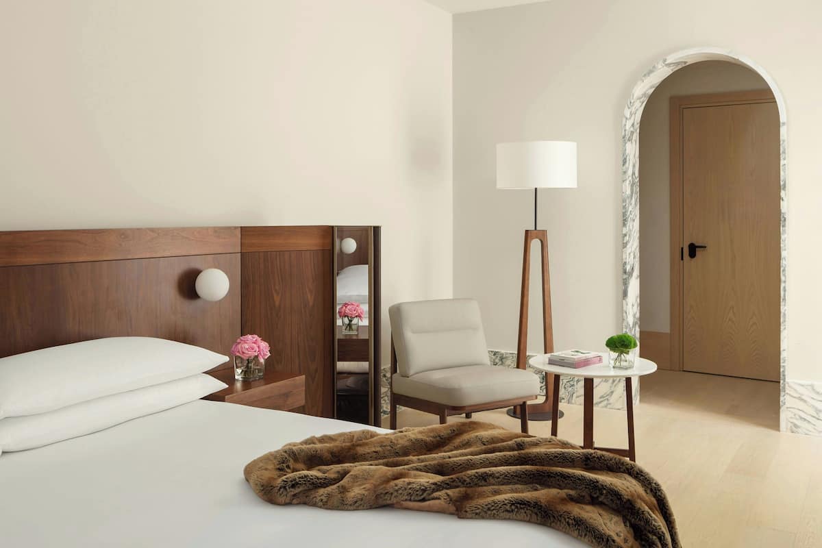 A hotel room with a bed, a chair, a small table with a lamp, and a door.