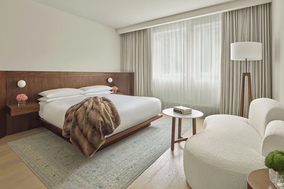 A modern hotel room with a large bed, a bedside table with a lamp, a chair, and a small table with a book.