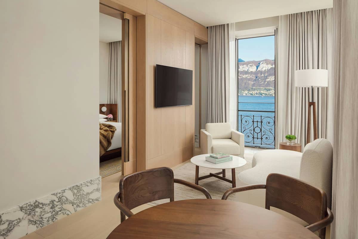 A hotel room with a balcony, a TV mounted on the wall, a dining table with chairs, and a view of a lake and mountains.