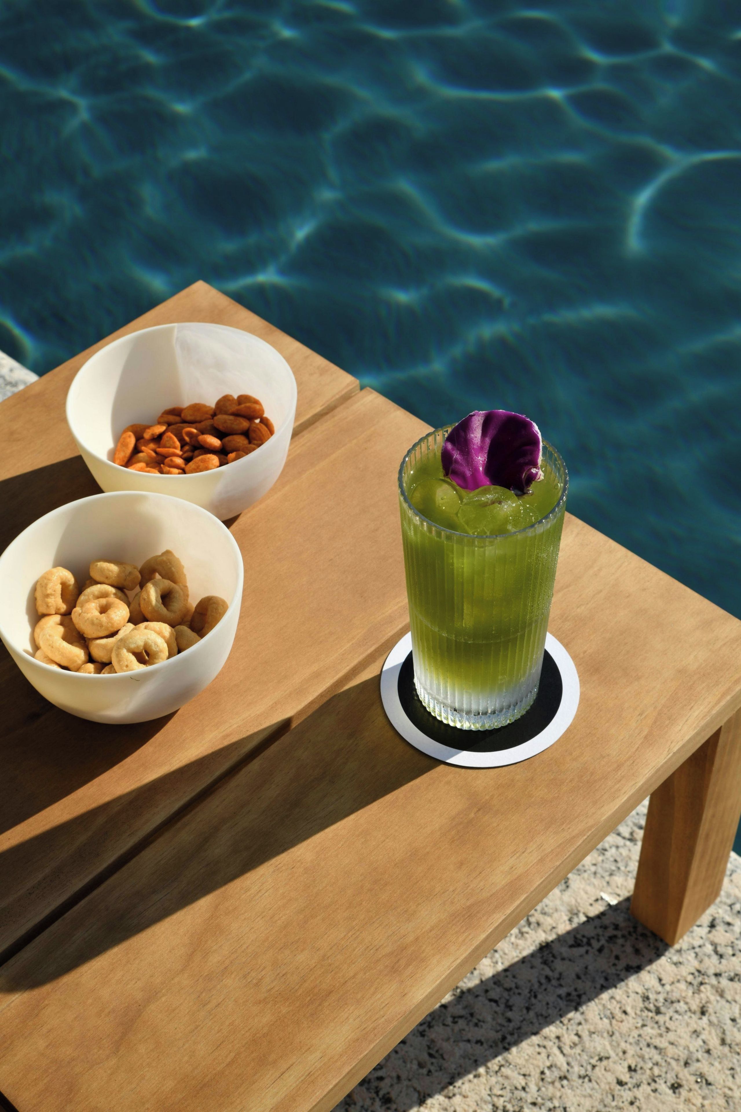 A wooden table with two bowls of snacks and a tall glass of green beverage with a purple flower garnish, set against a poolside background.