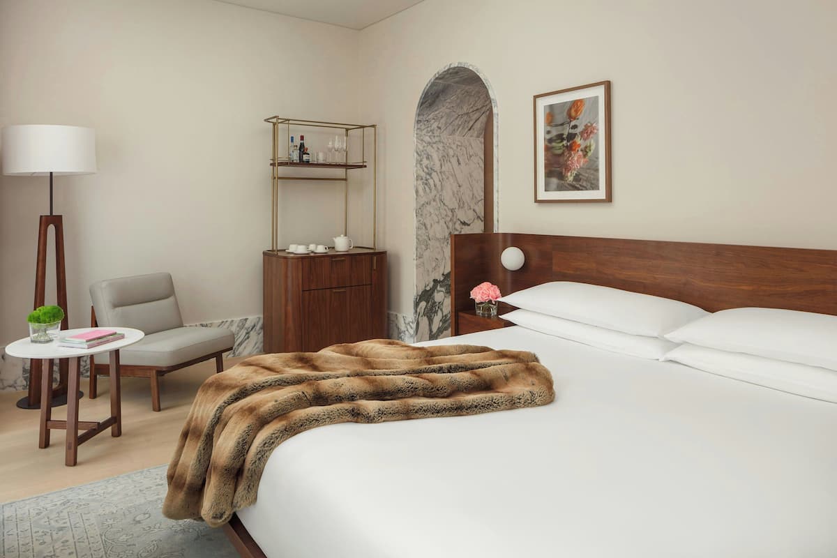 A hotel room with a large bed, a wooden nightstand, a chair, a small table with a lamp, a marble wall, and a shelf with toiletries.