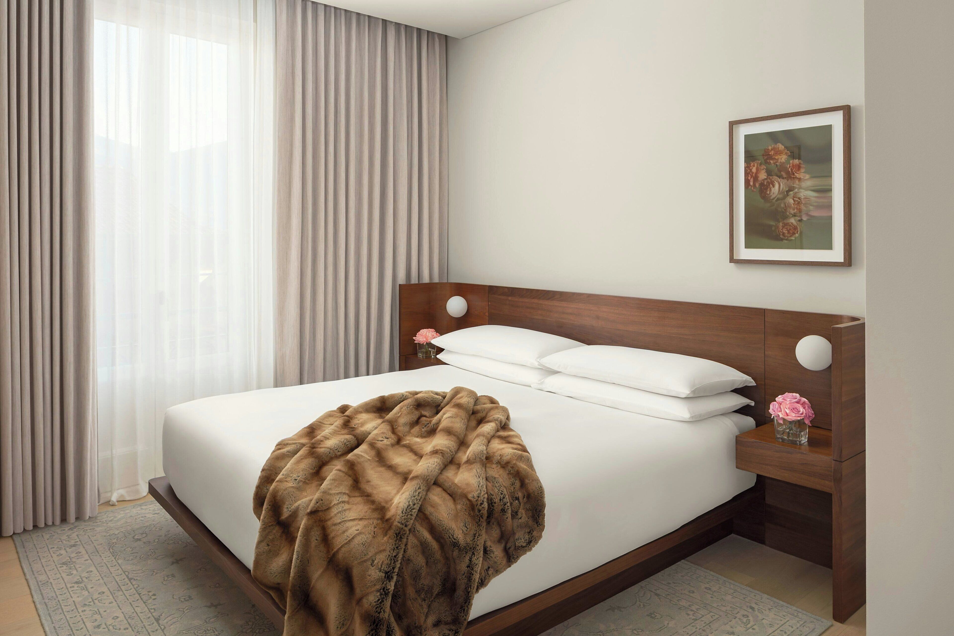 A neatly made bed with a white comforter and a brown throw blanket, a wooden headboard, a bedside table with a vase of pink flowers, and a framed picture on the wall.