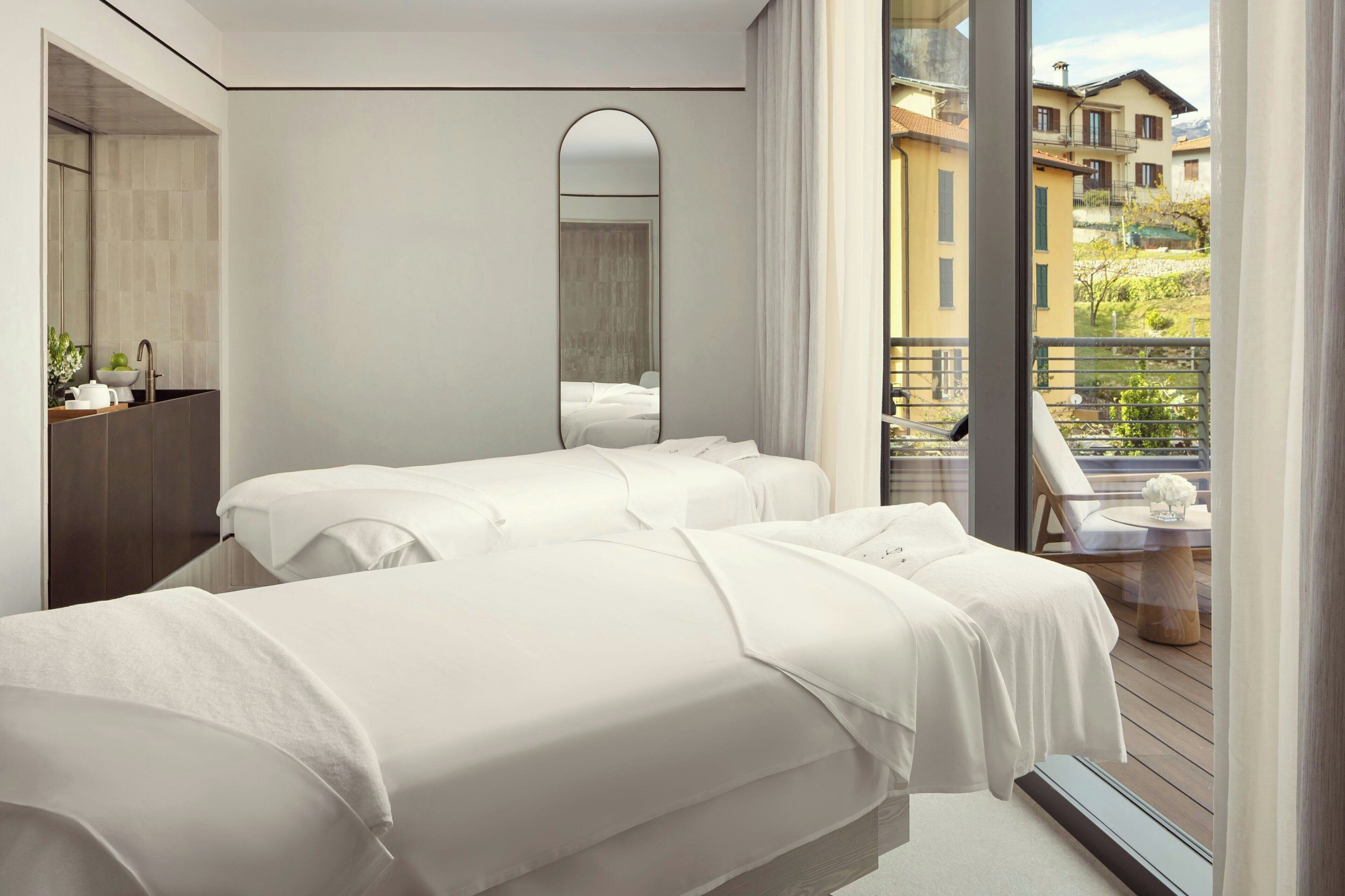 Quarto de hotel moderno com duas camas, um espelho grande, uma varanda com mesa e cadeiras, e vista para edifícios.