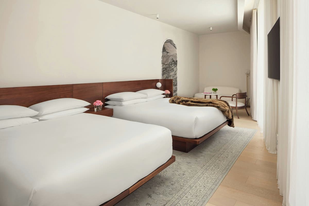 A modern hotel room with two beds, a small table with a plant, a chair, and a TV mounted on the wall.