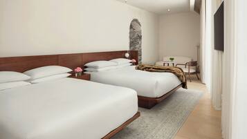 A modern hotel room with two beds, a small table with a plant, a chair, and a TV mounted on the wall.