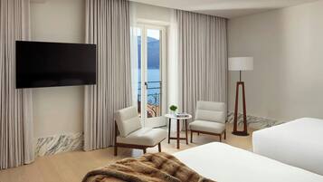 A hotel room with a bed, a TV mounted on the wall, a balcony with curtains, two chairs, a small table with a lamp, and a view of the sea and mountains.